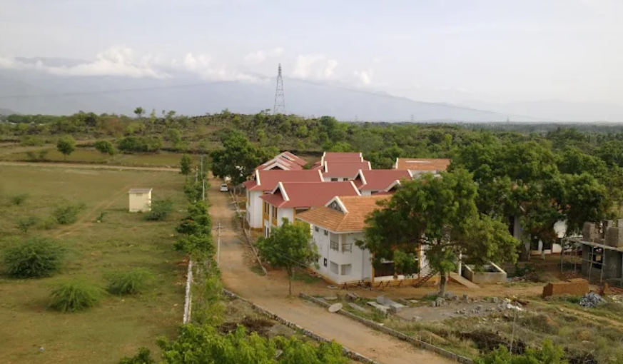 Aerial overview of Melur Meadows retirement village, showing tiled-roof cottages surrounded by green trees, open landscapes, and hills under an overcast sky.