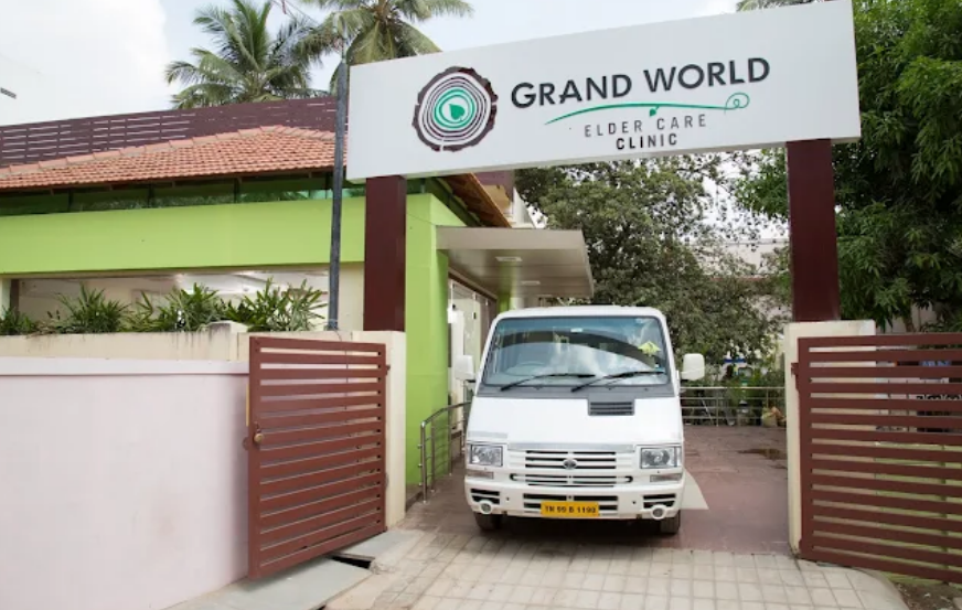 Exterior view of the Grand World Elder Care Clinic facility entrance in R.S. Puram, featuring a prominent signboard, front gate, and tiled roof surrounded by palm trees.